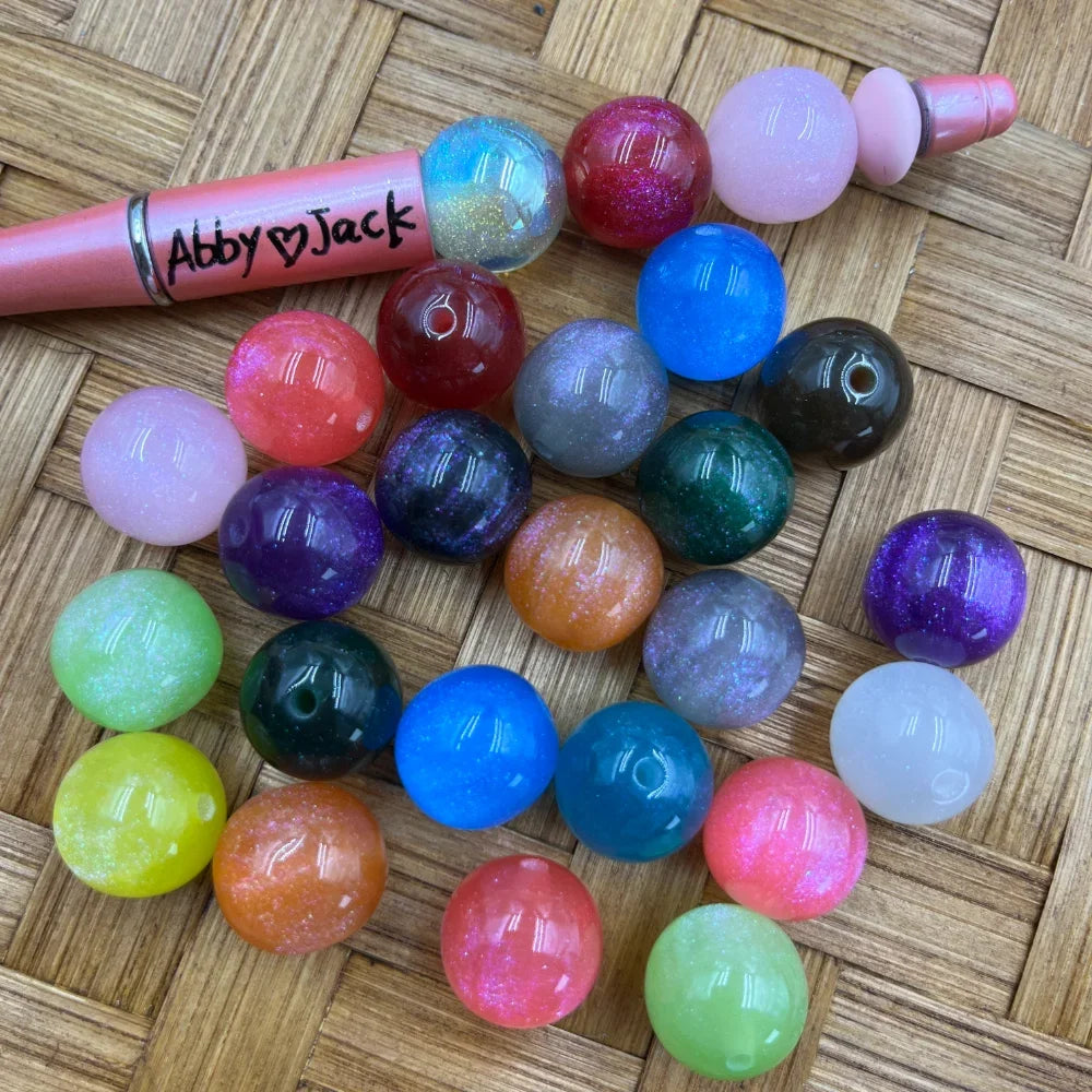 Assorted 16MM glitter galaxy acrylic beads in multiple colors including pink, blue, purple, teal, orange, green, and clear, displayed next to a pink beadable pen on a wicker surface.