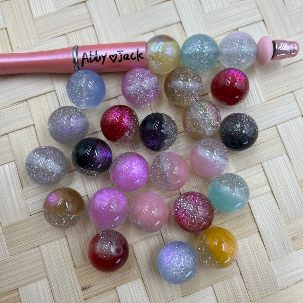 16MM glitter galaxy acrylic beads in assorted colors including pink, red, black, purple, teal, gold, and clear with sparkle finish, displayed next to a pink beadable pen on a wicker surface.