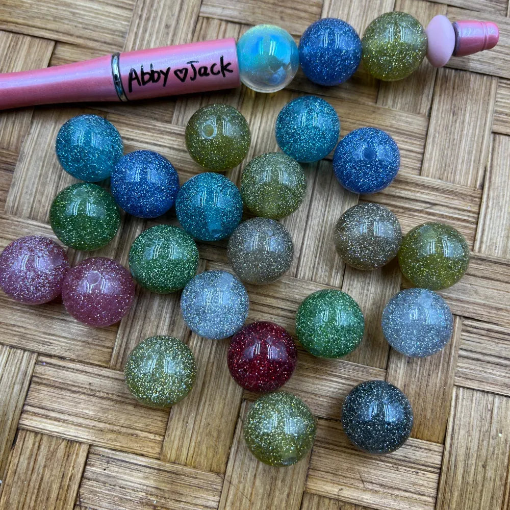 16MM multicolor glitter galaxy acrylic beads in blue, green, pink, red, silver, and teal sparkle finishes displayed on woven surface beside a pink beadable pen labeled Abby and Jack.