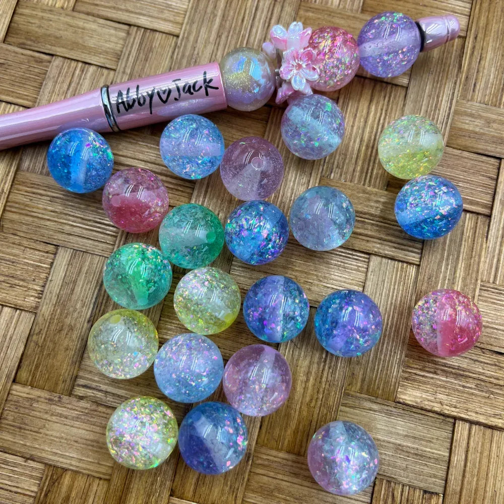 16MM pastel rainbow glitter galaxy crackle acrylic beads in assorted colors including blue, purple, pink, green, and yellow with holographic sparkle flakes, displayed on a woven surface next to a pink beadable pen.