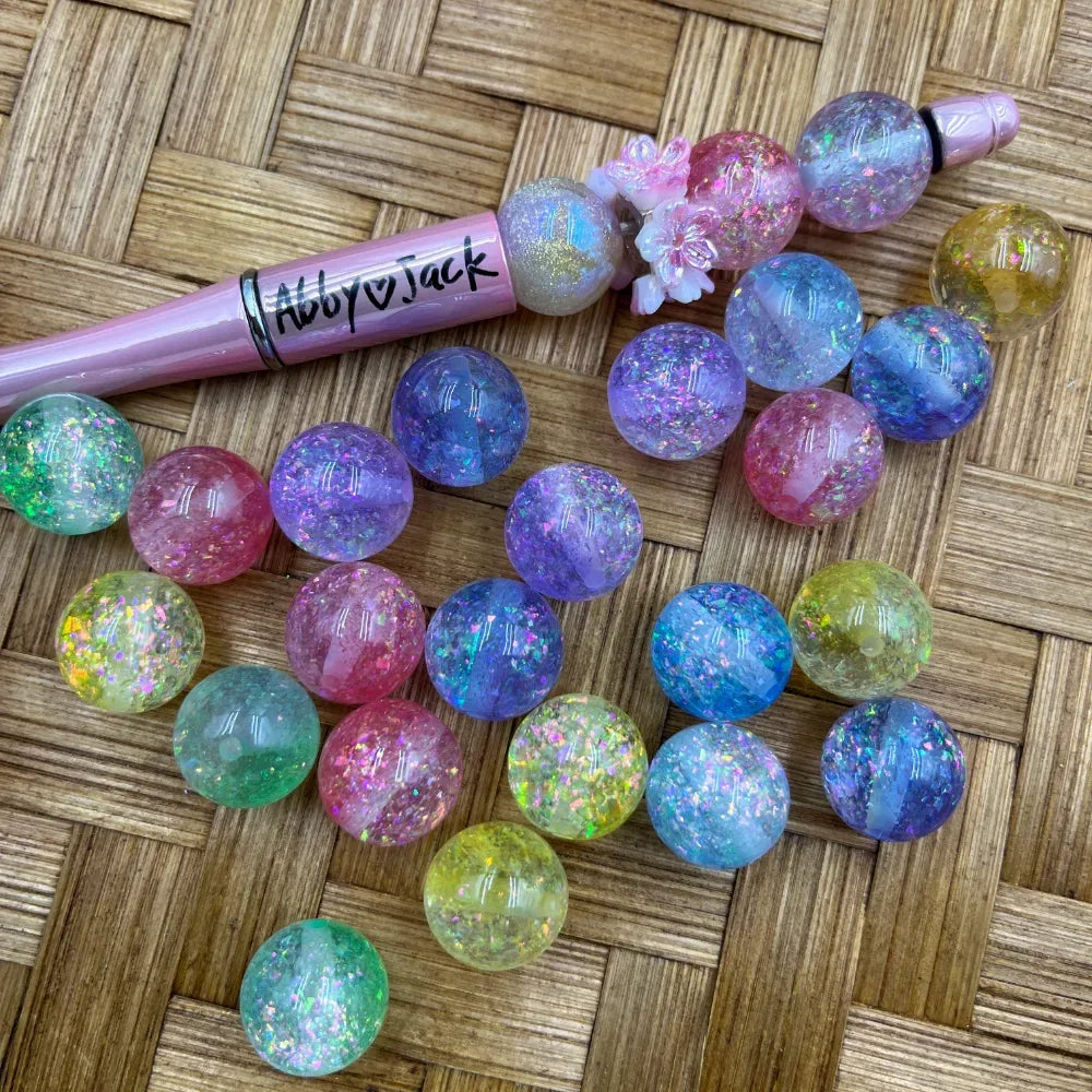 16MM pastel rainbow glitter galaxy crackle acrylic beads in pink, purple, blue, green, and yellow scattered on wicker surface next to a beadable pen.