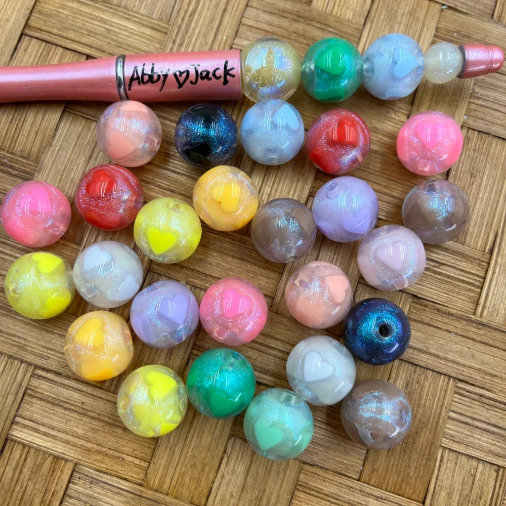 16MM round acrylic beads with glitter finish and embedded heart details in assorted pastel and vibrant colors displayed on a woven surface next to a beadable pen labeled Abby Jack.