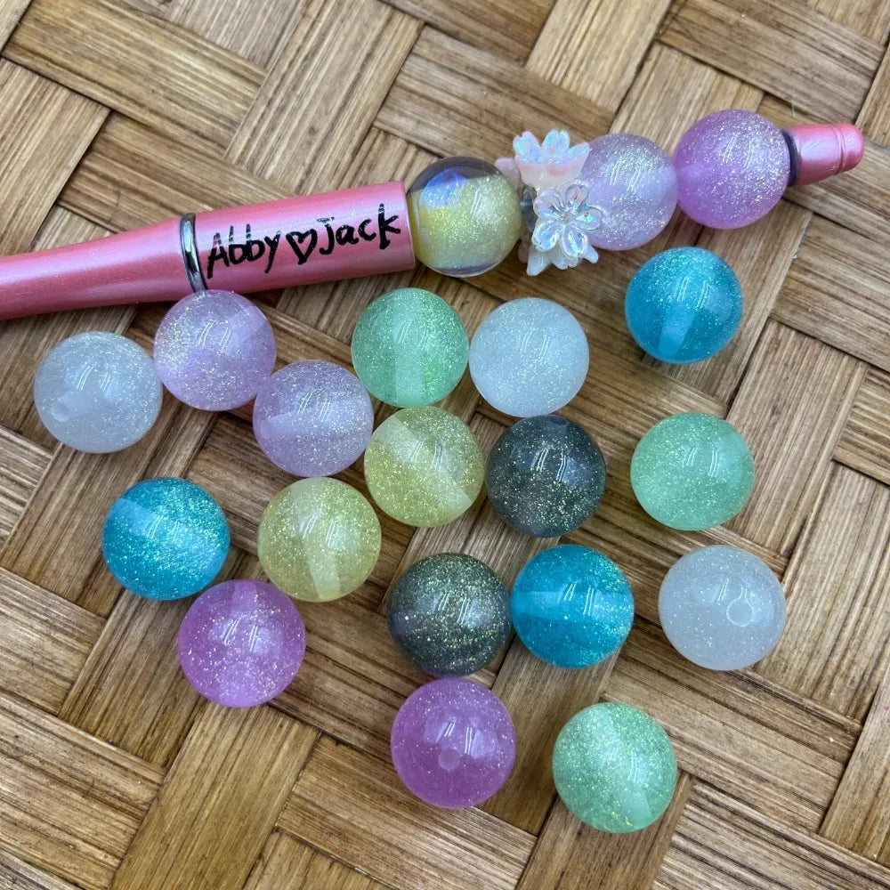 16MM glitter pastel acrylic beads in assorted colors including pink, purple, teal, green, yellow, gray, and white with sparkle finish, displayed next to a pink beadable pen on a wicker surface.