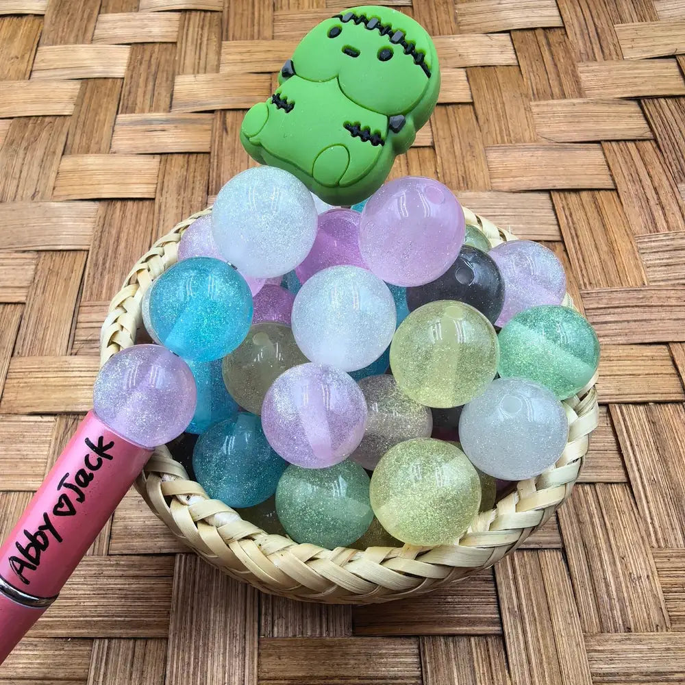 16MM glitter pastel iridescent acrylic beads in a wicker basket with mint green, lavender, blue, clear, and yellow-green sparkle finishes, shown with a beadable pen and dinosaur topper.