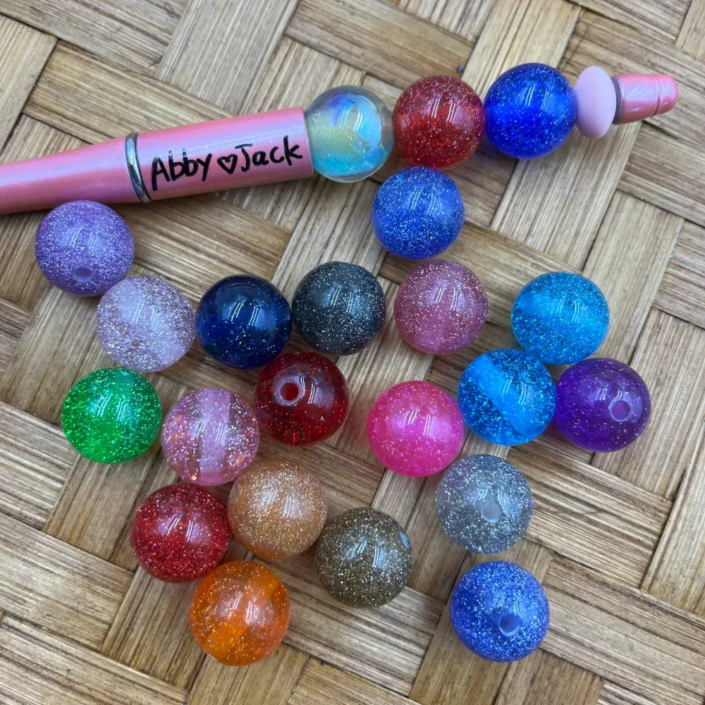 16MM glitter sparkle acrylic beads in assorted colors including blue, pink, purple, red, green, orange, and gold displayed next to a beadable pen on a wicker surface.