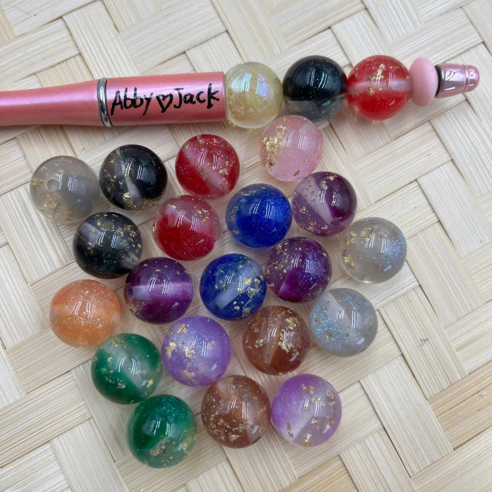 16MM gold foil galaxy marble acrylic beads in assorted colors including black, red, blue, purple, green, pink, and clear, displayed next to a beadable pen on a woven surface.