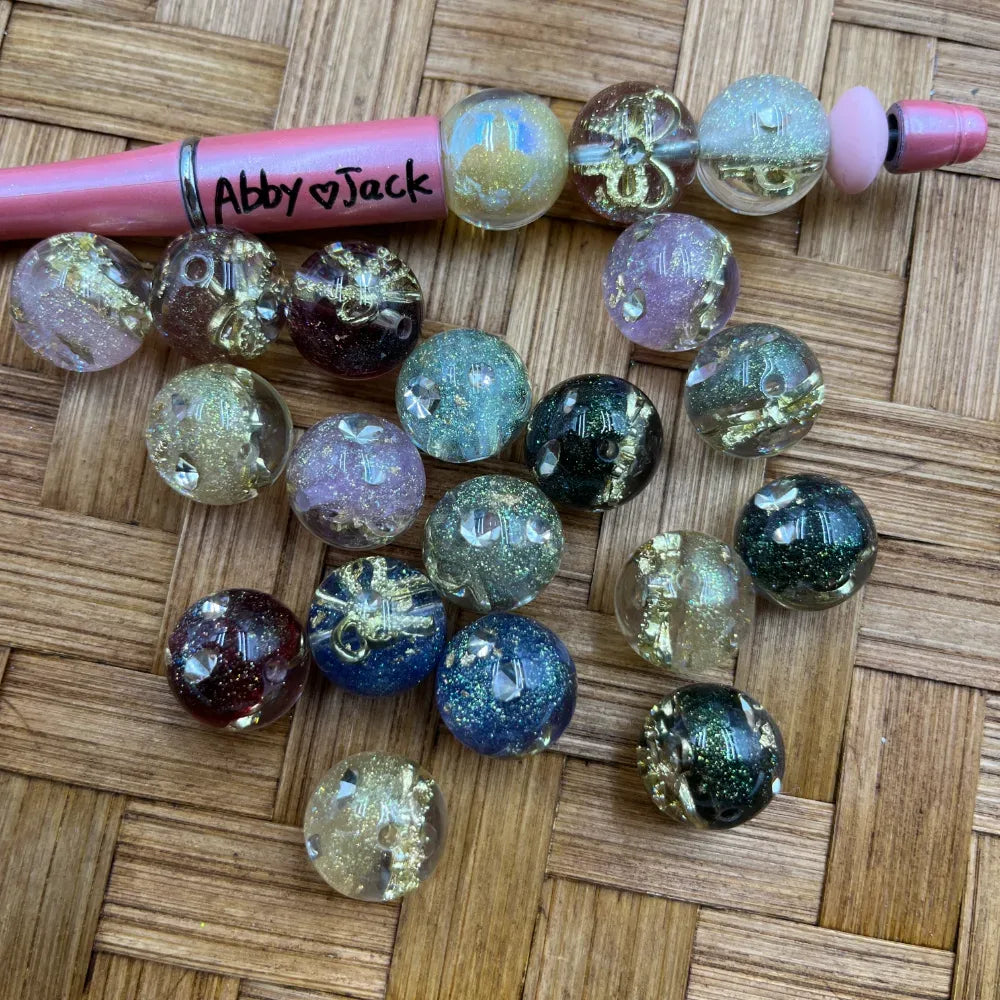 16MM round acrylic beads with gold foil and glitter galaxy effect in mixed colors including teal, navy, burgundy, champagne, lavender, and clear displayed next to a pink beadable pen on a wicker surface.