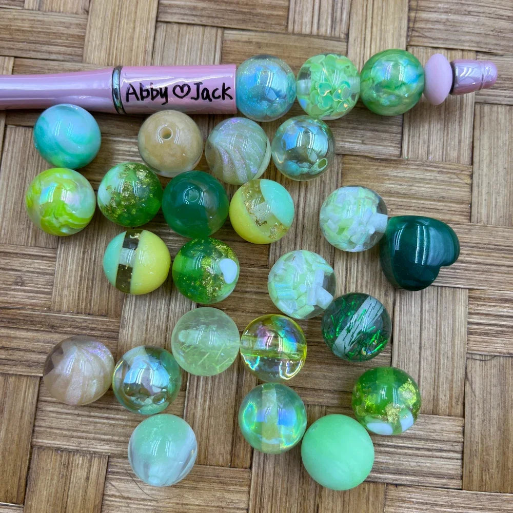 Assorted 16MM green mix acrylic beads in lime, mint, emerald, and iridescent finishes displayed on a woven surface next to a pink beadable pen labeled Abby and Jack.