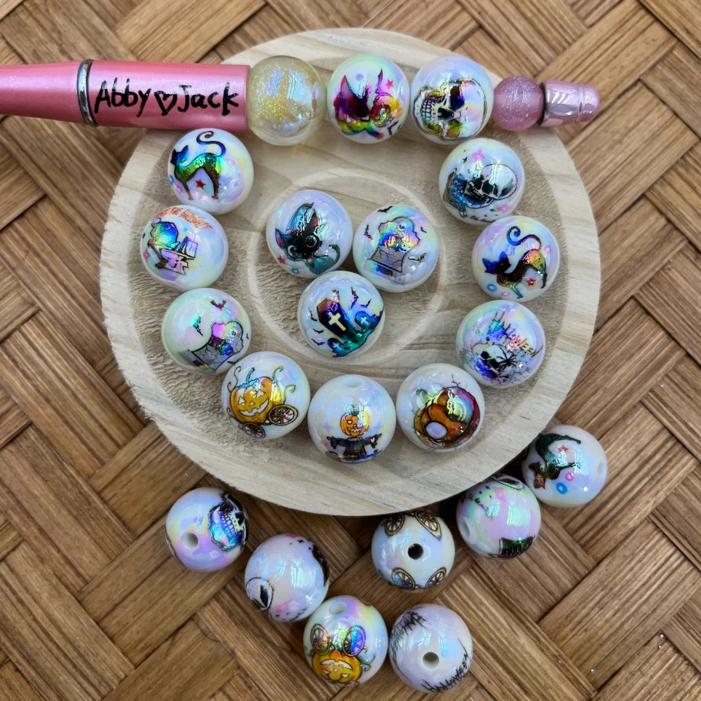 16MM iridescent white acrylic beads with colorful Halloween prints including pumpkins, skulls, black cats, coffins, and scarecrows arranged on a wooden display tray with a pink beadable pen.