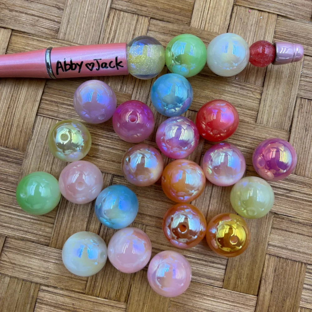 16MM iridescent AB finish round acrylic beads in assorted pastel and vibrant colors including pink, green, white, orange, red, and blue, displayed next to a beadable pen on a wicker surface.