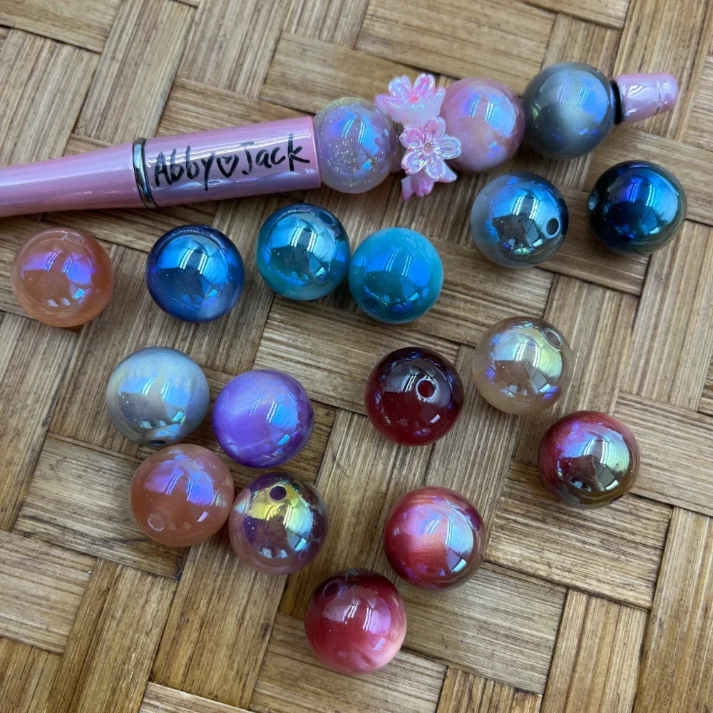16MM iridescent aurora AB acrylic beads in assorted colors including blue, teal, purple, pink, red, and gray with holographic shimmer finish, displayed next to a beadable pen on a wicker surface.