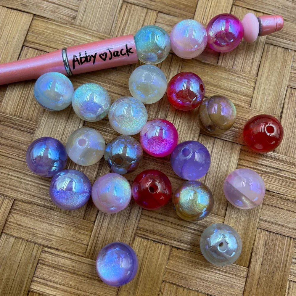 16MM iridescent aurora AB acrylic beads in mixed colors including purple, pink, red, clear, and white with rainbow shimmer finish, shown with a beadable pen on a woven surface.