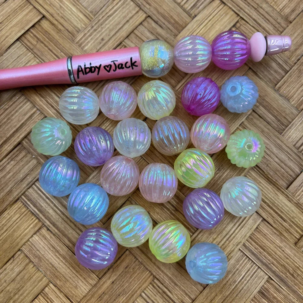 Iridescent aurora AB ribbed acrylic beads in pastel mixed colors including pink, purple, blue, green and white, displayed next to a pink beadable pen on a woven surface.