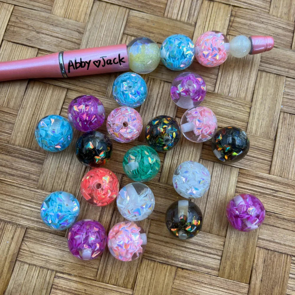 16MM iridescent confetti glitter acrylic beads in assorted colors including teal, pink, purple, black, coral, white and blue displayed next to a beadable pen on a woven surface