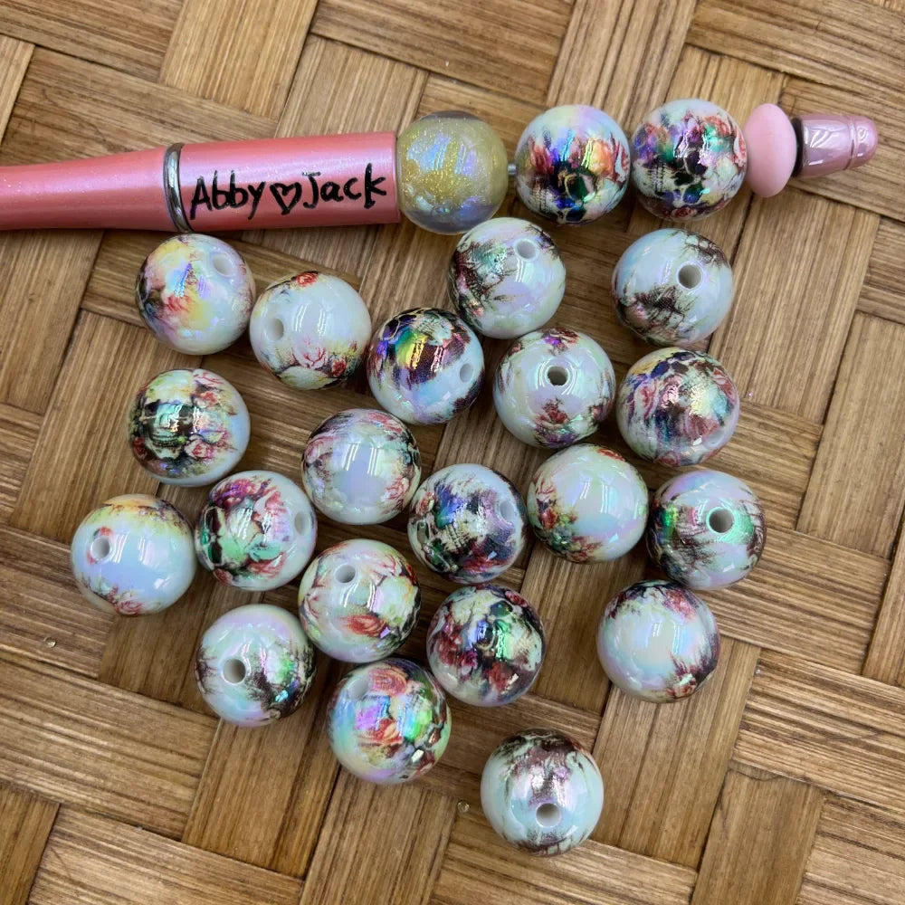 16MM iridescent floral marble acrylic beads in white with multicolor holographic shimmer and rose floral print, shown with a beadable pen on a wicker surface.