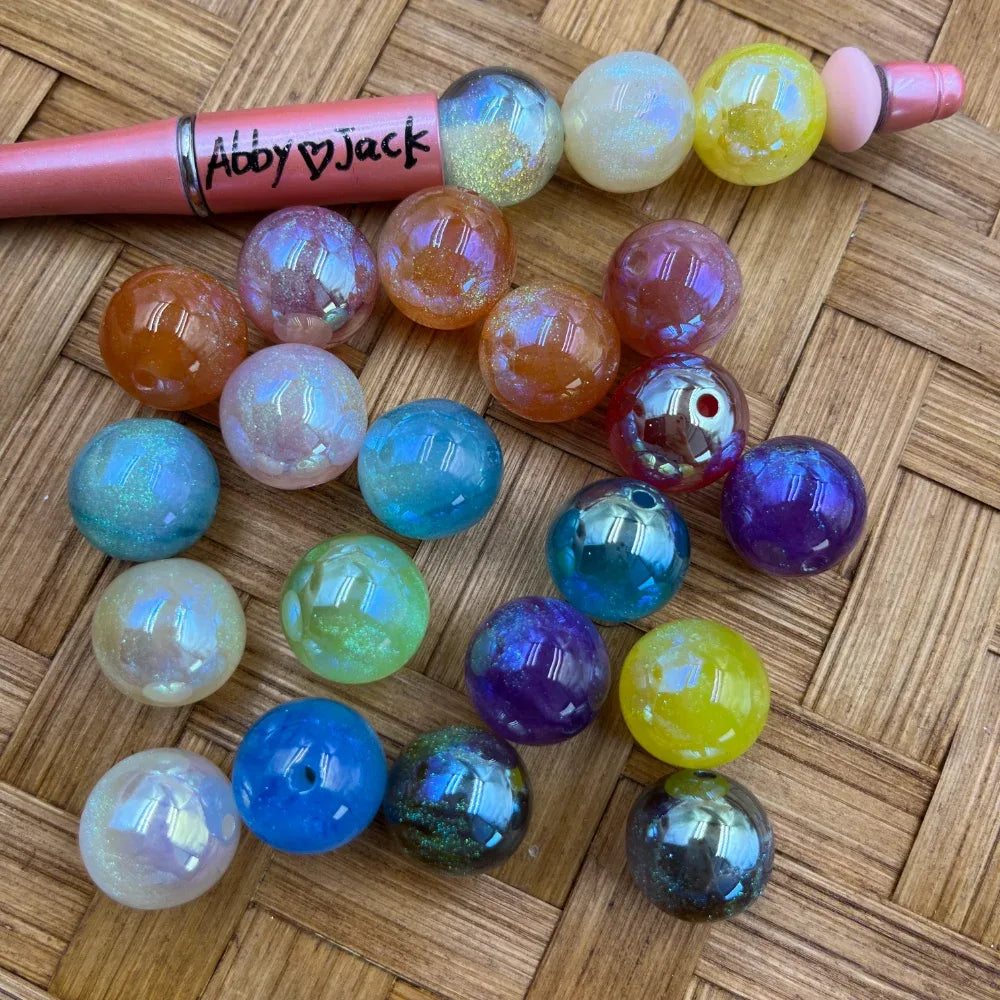 16MM iridescent galaxy glitter acrylic beads in assorted colors including blue, purple, yellow, orange, teal, and white with a shimmer AB finish, displayed on a wicker surface next to a pink beadable pen.