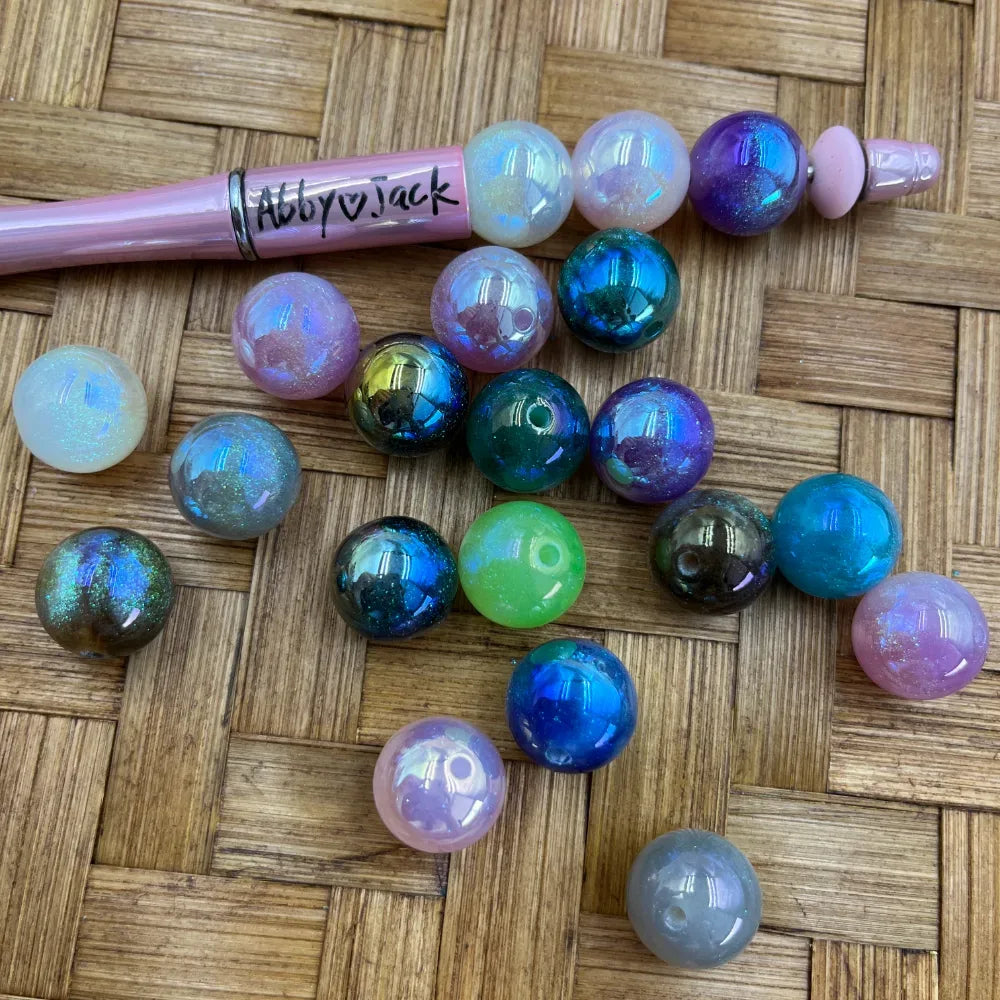 16MM iridescent galaxy acrylic beads in mixed holographic colors including blue, green, purple, pink, and opal white, displayed next to a pink beadable pen on a wicker surface.