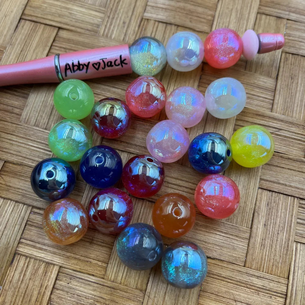 16MM iridescent glitter aurora acrylic beads in mixed colors including blue, red, green, orange, yellow, pink, and clear, displayed next to a pink beadable pen on a wicker surface.