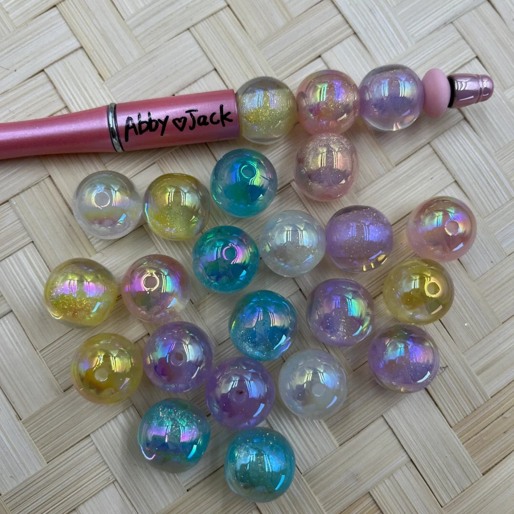 16MM iridescent pastel glitter galaxy acrylic beads in teal, purple, pink, yellow, and clear aurora finish displayed with a beadable pen on wicker surface.