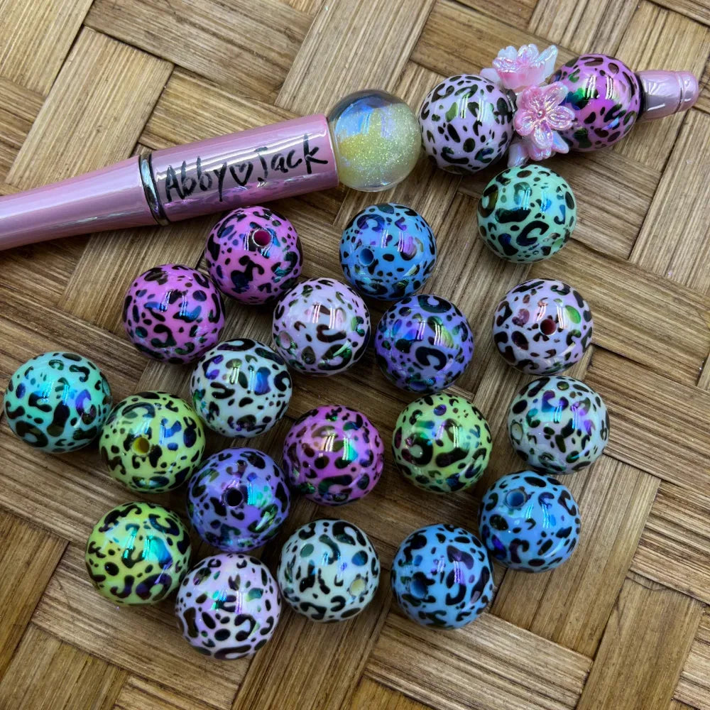 16MM iridescent leopard print acrylic beads in assorted colors including pink, teal, yellow, purple, and blue with black spots, displayed on woven surface next to a beadable pen.