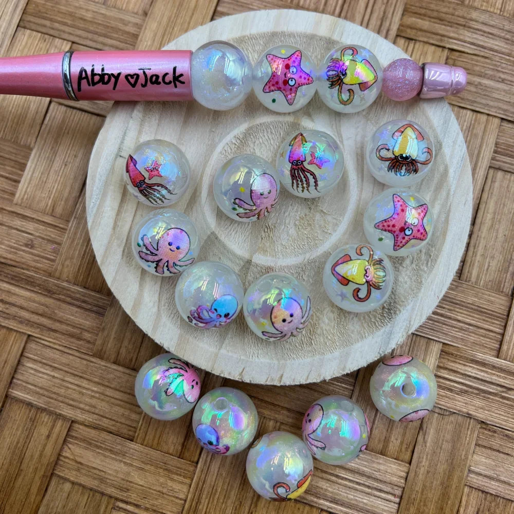 16MM iridescent acrylic beads with cute ocean sea creature designs including octopus, starfish, jellyfish, and squid, displayed on a wooden tray with a pink beadable pen.