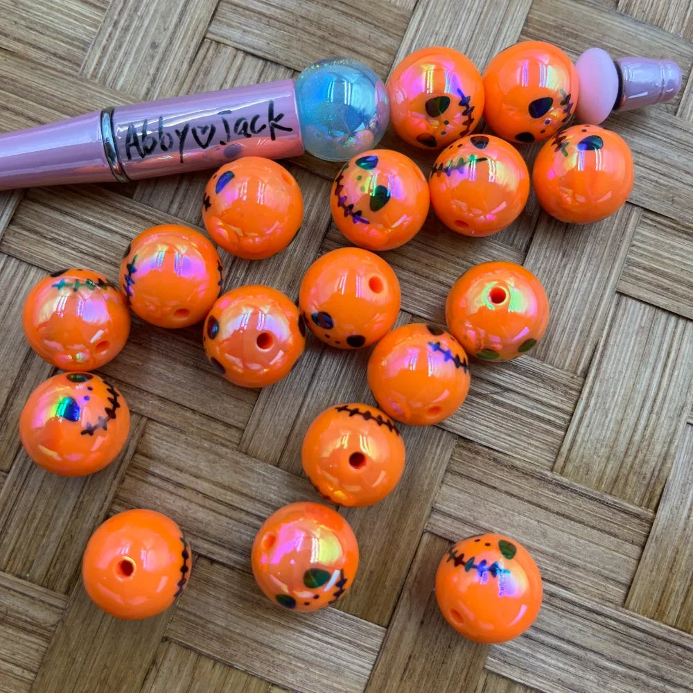 16MM orange iridescent jack-o-lantern acrylic beads with pumpkin face design displayed next to a pink beadable pen on a wicker surface