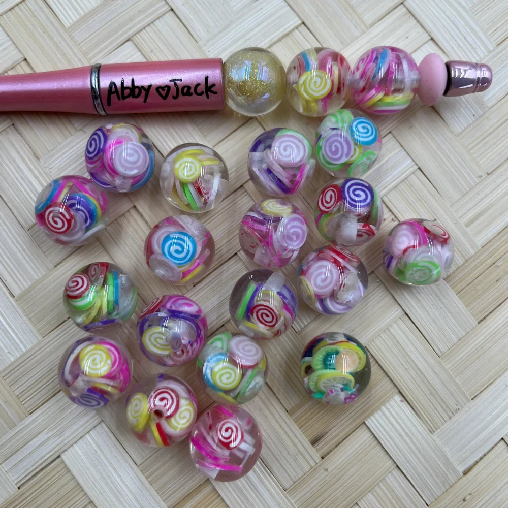 16MM clear acrylic round beads with colorful lollipop swirl candy designs in pink, purple, yellow, red and green, displayed next to a pink beadable pen on a woven surface.