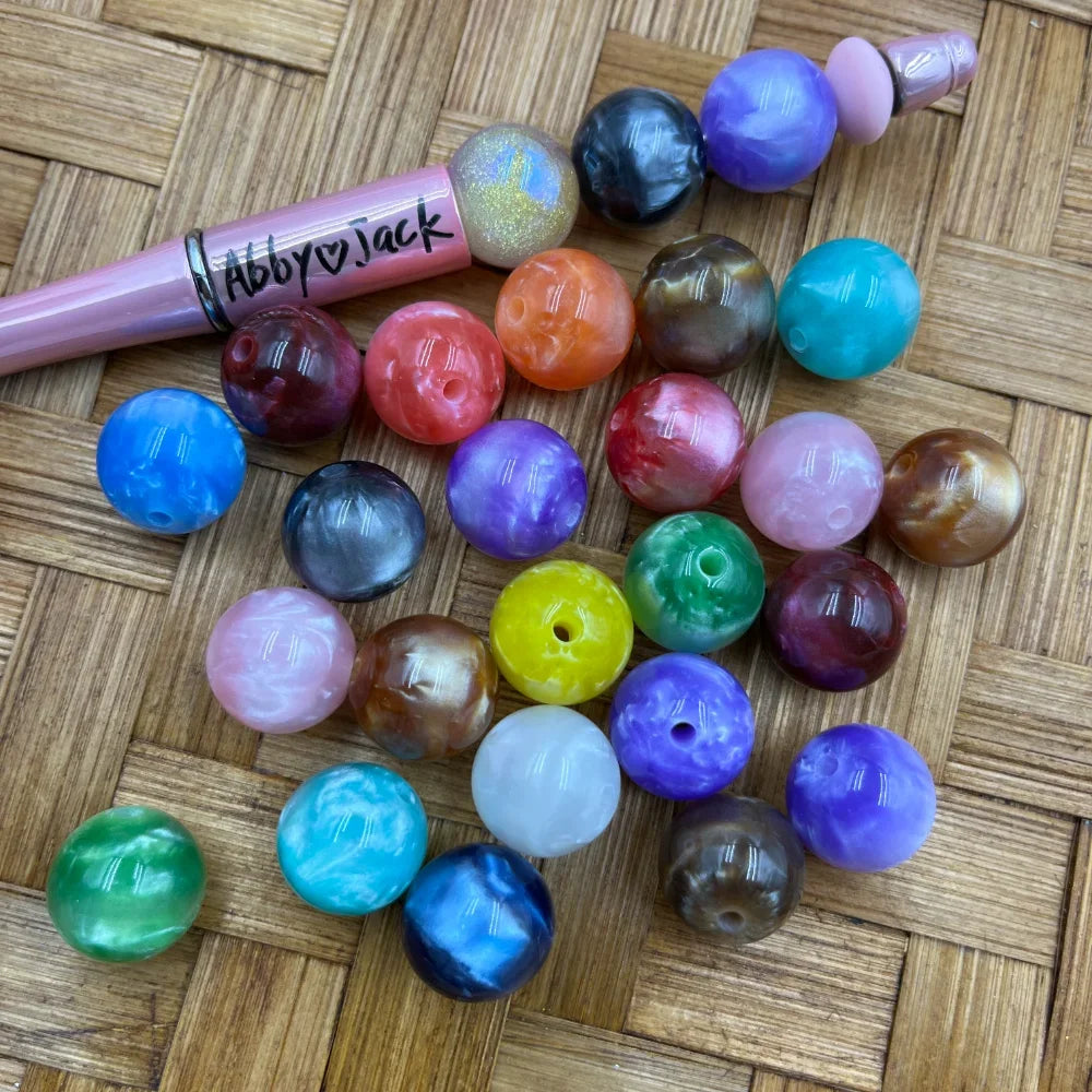 16MM marble swirl acrylic beads in assorted colors including blue, purple, pink, green, teal, yellow, brown, and gray displayed on a woven surface next to a pink beadable pen labeled Abby Jack.