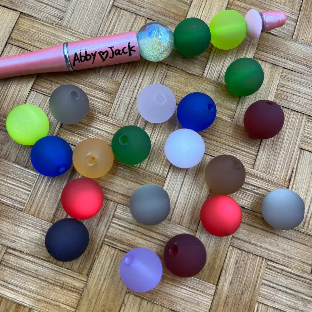 16MM matte frosted acrylic beads in assorted colors including red, blue, green, gray, white, navy, and maroon displayed on a woven surface next to a pink beadable pen labeled Abby Jack.