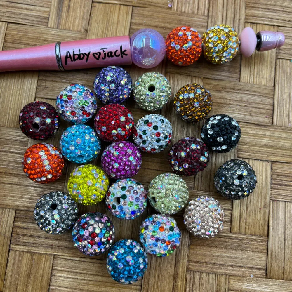 16MM mixed color clay rhinestone beads in assorted colors including red, blue, orange, yellow, purple, black, green, and multicolor disco styles displayed on a wicker surface next to a beadable pen.