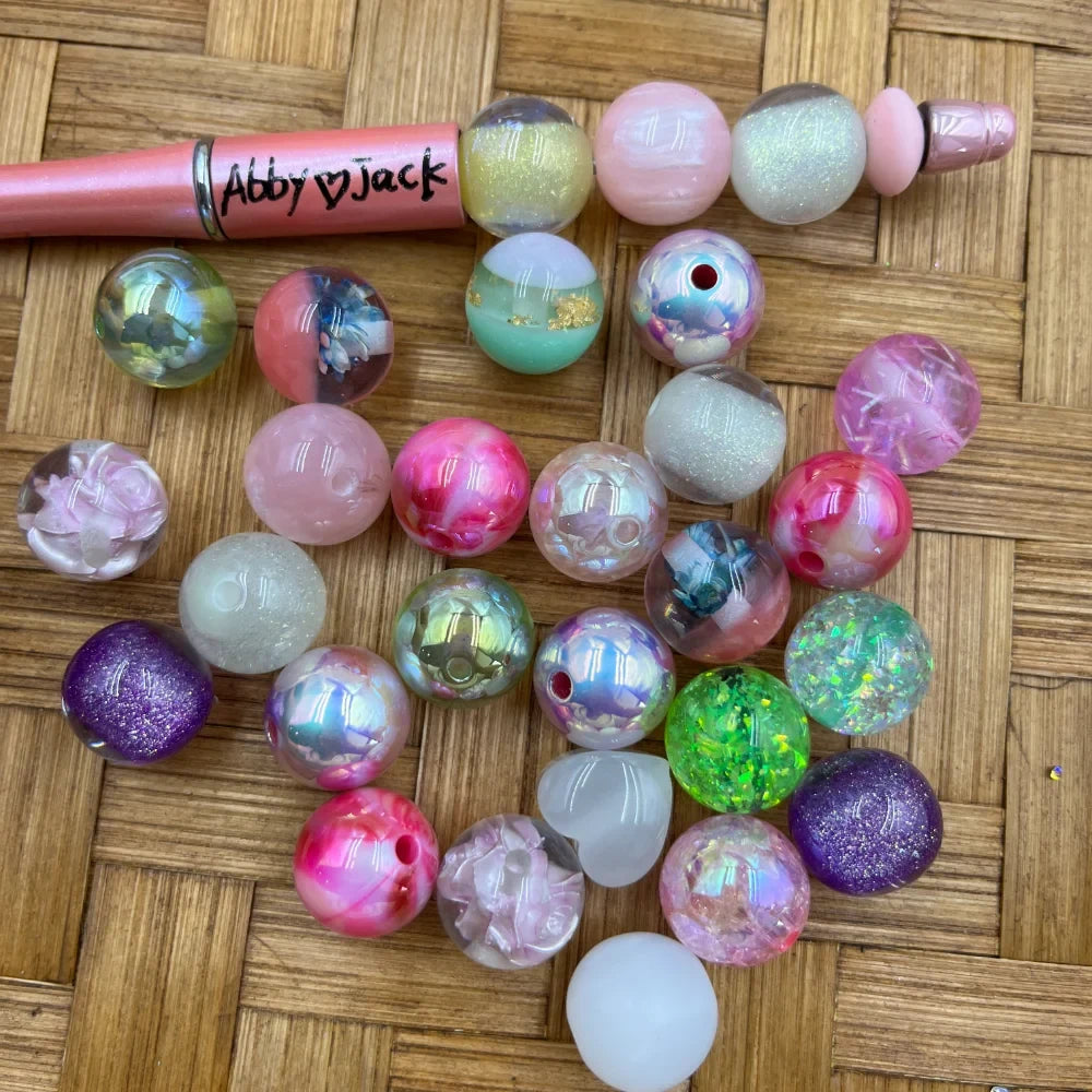 Assorted 16MM acrylic beads in galaxy glitter, iridescent, marble swirl, and chunky confetti finishes in pink, purple, green, and white, displayed beside a beadable pen on a wicker surface.