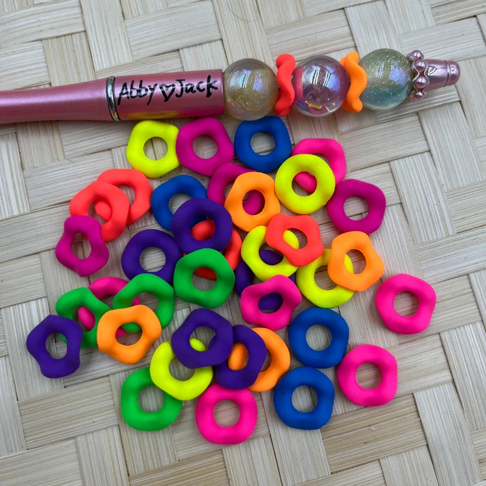 16MM neon multicolor flower-shaped ring acrylic spacer beads in pink, orange, green, purple, blue, and yellow scattered on a woven surface next to a decorated beadable pen.