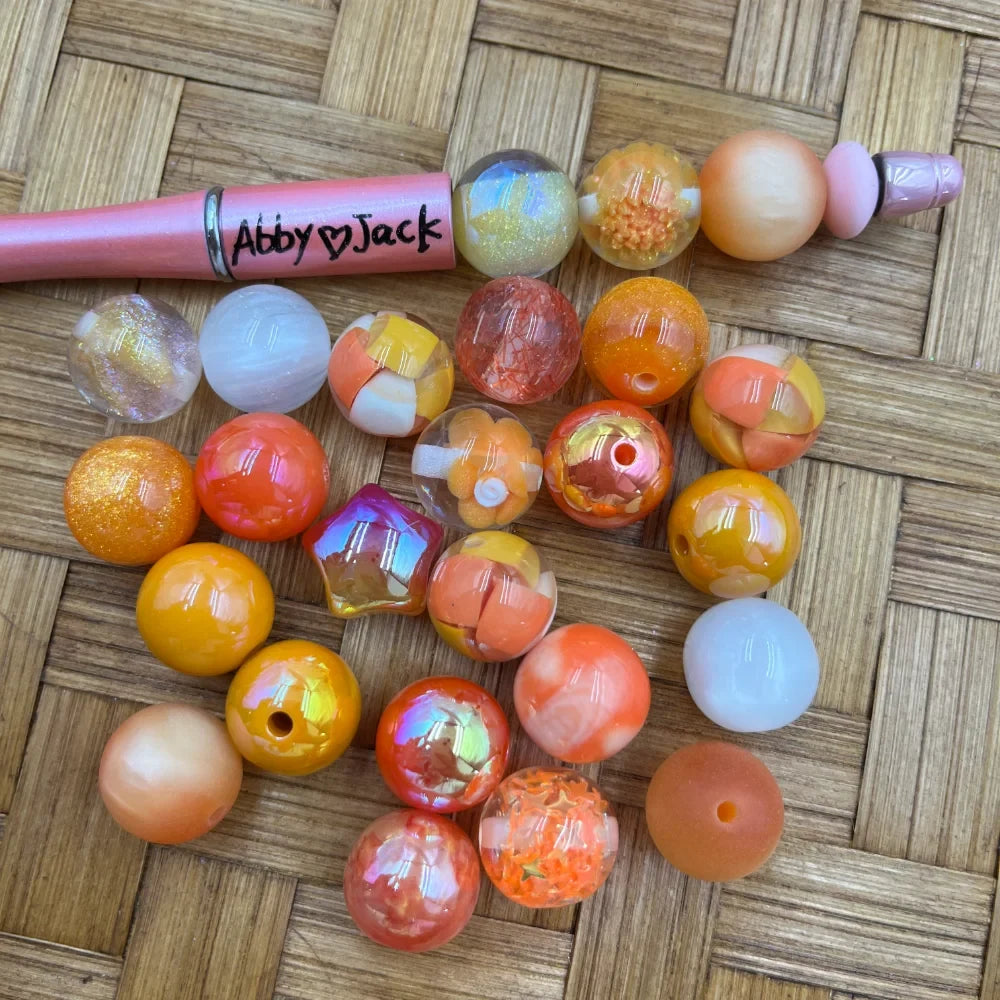 Assortment of 16MM orange, yellow, and peach acrylic beads in various finishes including glitter, iridescent, marble, and solid, displayed next to a pink beadable pen on a wicker surface.