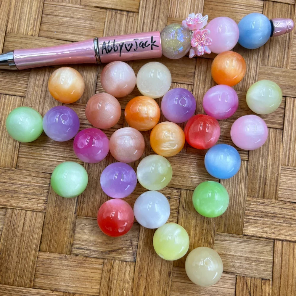 16MM pastel gumball acrylic beads in assorted candy colors including pink, purple, green, blue, orange, red, and yellow arranged on a wicker surface next to a pink beadable pen.