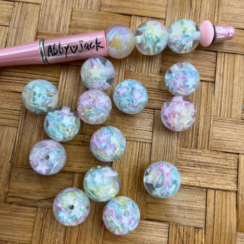 16MM round acrylic beads filled with pastel iridescent star confetti in pink, blue, yellow, and mint, displayed on a woven surface next to a pink beadable pen.
