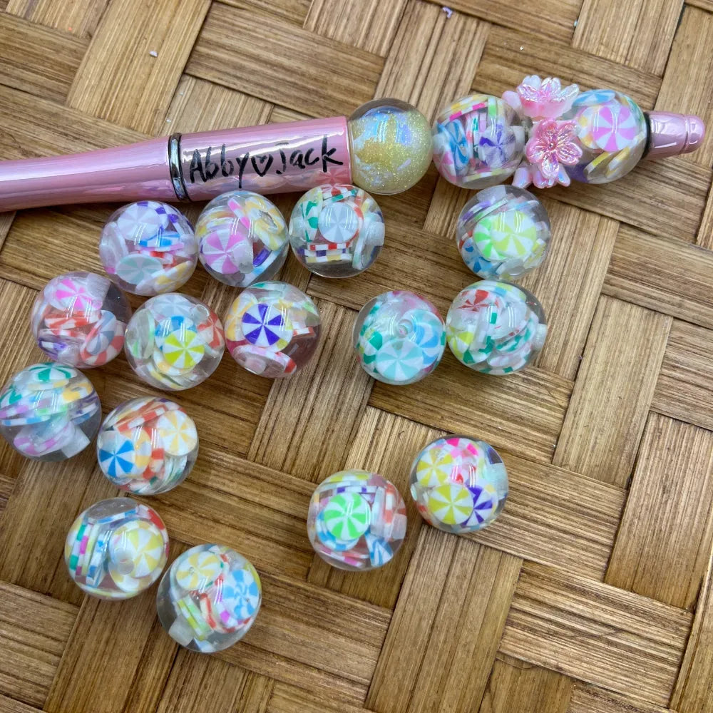 16MM clear acrylic round beads filled with colorful peppermint candy confetti slices in assorted colors, displayed next to a pink beadable pen on a wicker surface.