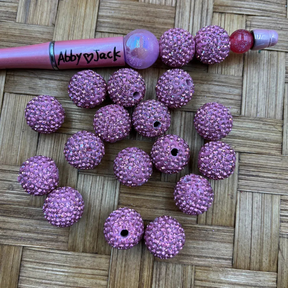 16MM pink clay rhinestone beads with iridescent crystal embellishments displayed on a wicker surface next to a beaded pen, showing drilled holes for jewelry making.