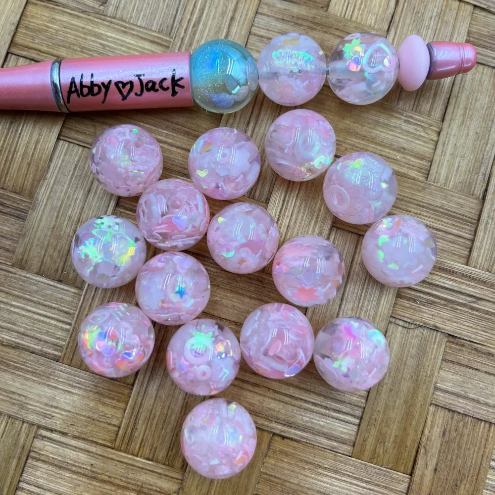 16MM pink confetti glitter acrylic beads with iridescent holographic shimmer, shown loose and strung on a beadable pen on a woven bamboo surface.