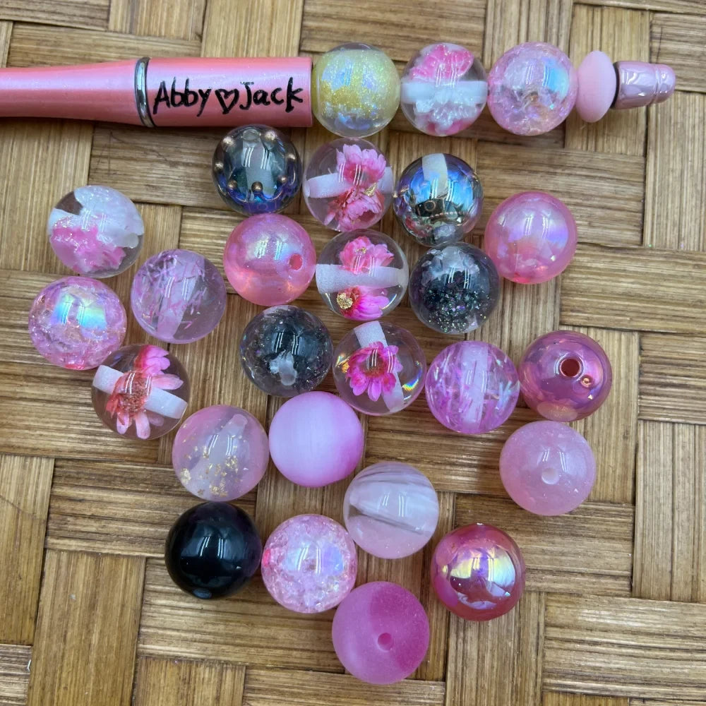 16MM pink floral galaxy glitter acrylic beads in assorted styles including iridescent, crackle, black, and flower encased designs displayed on woven surface next to a pink beadable pen labeled Abby Jack.