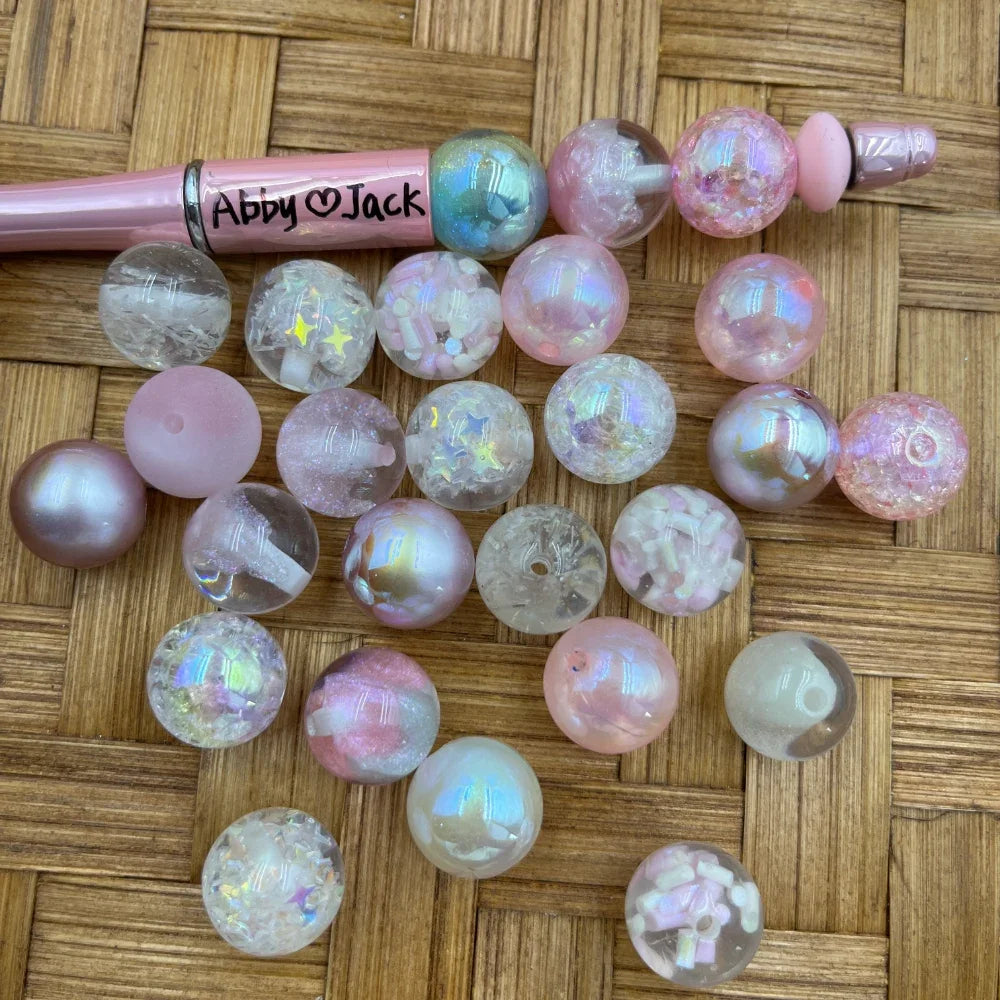 16MM pink iridescent acrylic beads in assorted finishes including galaxy glitter, confetti, pearl, and crackle effects displayed next to a pink beadable pen on a woven bamboo surface.