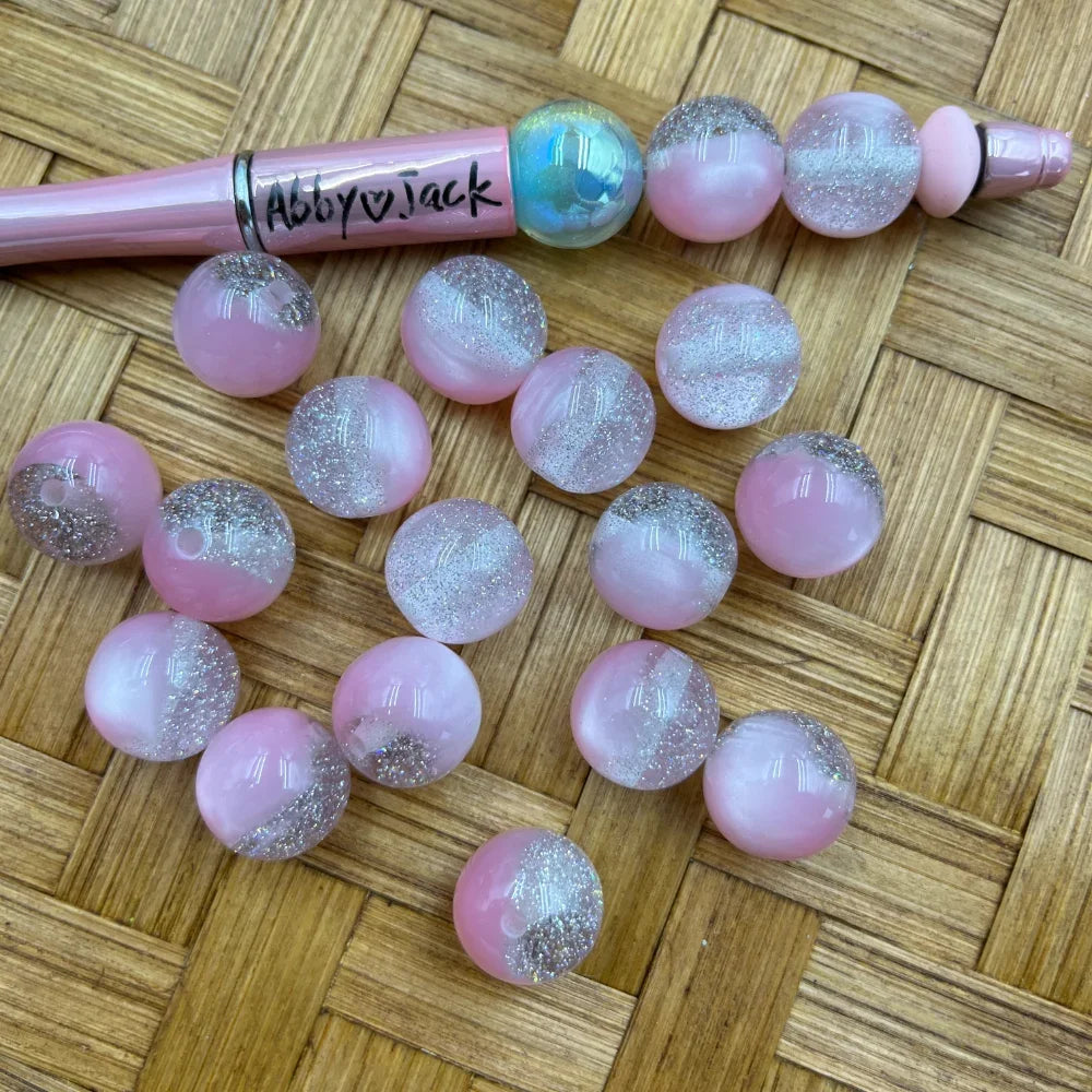 16MM pink and silver glitter ombre two-tone acrylic beads displayed on a wicker surface next to a pink beadable pen, showing sparkly clear glitter top half and soft pink bottom half.