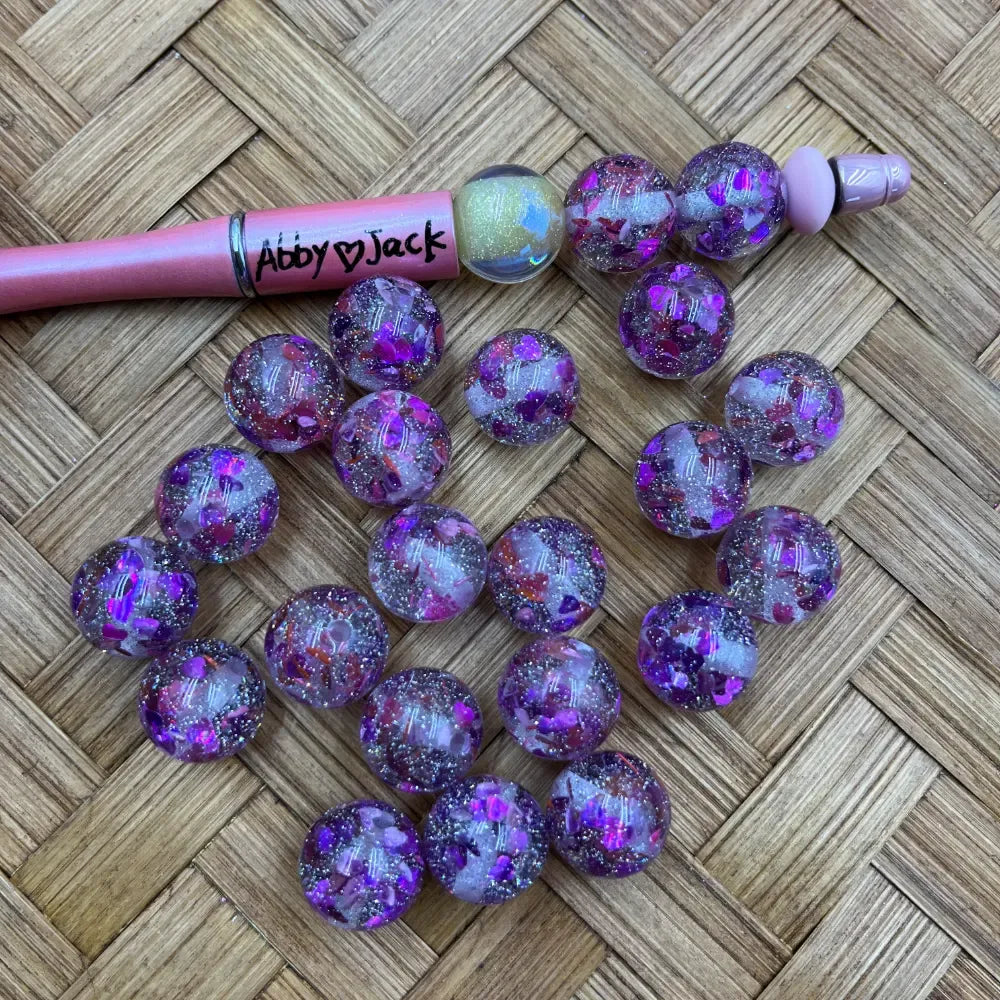 16MM purple glitter confetti acrylic beads with iridescent flakes and sparkle finish displayed on woven surface next to a pink beadable pen labeled Abby Jack