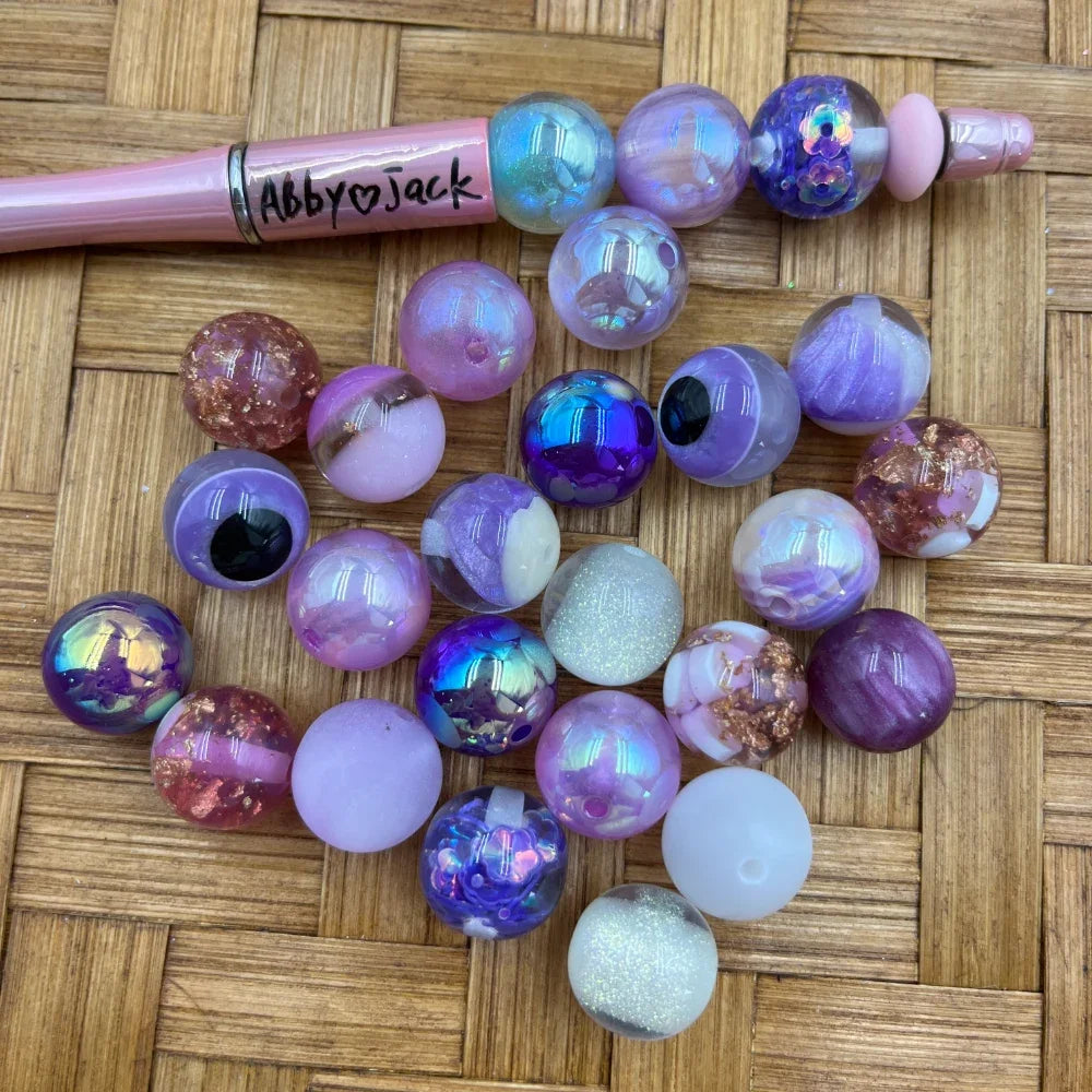 16MM purple iridescent galaxy acrylic beads in assorted lavender, violet, holographic, marble, and glitter finishes displayed next to a pink beadable pen on a wicker surface.