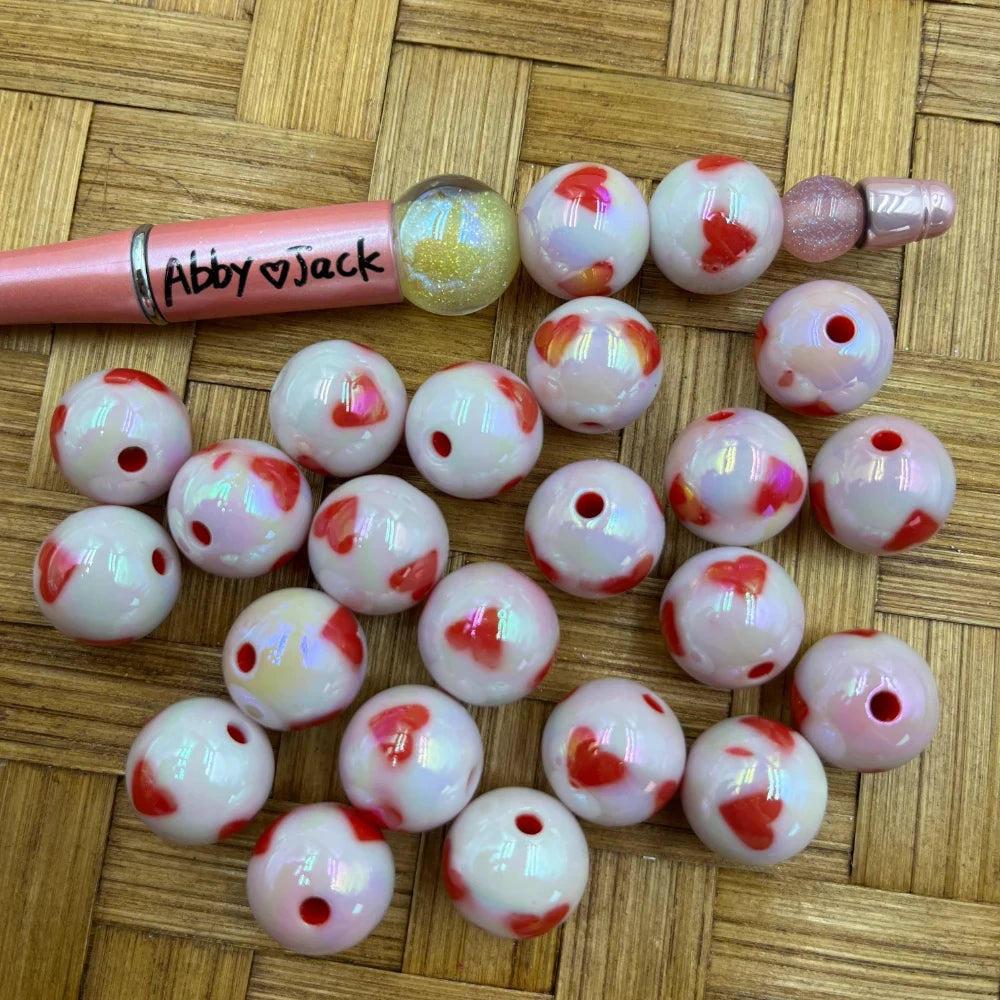 16MM white iridescent AB acrylic round beads with red heart pattern displayed on wicker surface next to a beadable pen, showing hole detail for DIY jewelry making.