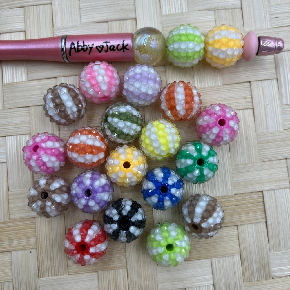Assorted 16MM round rhinestone beads with raised swirl stripe design in pink, purple, orange, green, blue, black, yellow, red, and brown, shown with a beadable pen.