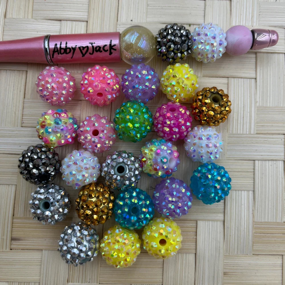 Assorted 16MM color mix AB pave rhinestone beads in pink, purple, blue, yellow, green, white, black, silver, and gold beside a beadable pen