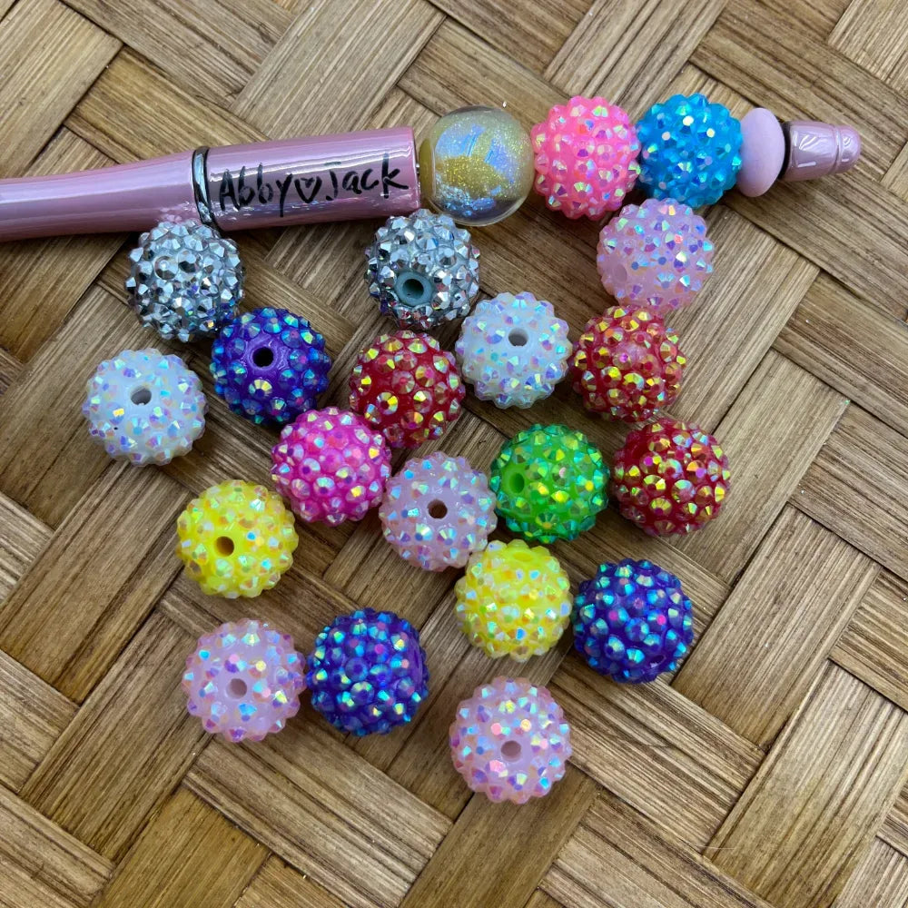 16MM multicolor rhinestone sparkle acrylic beads in assorted colors including pink, blue, yellow, green, red, purple, and white with iridescent AB finish, shown next to a beadable pen on a wicker surface.