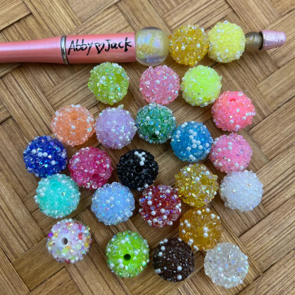 16MM mixed color rhinestone sugar beads in assorted colors including blue, pink, green, yellow, black, white, and orange displayed on woven surface next to a beadable pen