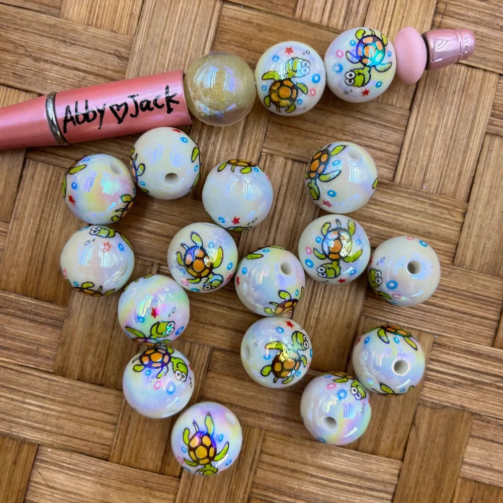 16MM white iridescent acrylic beads with colorful cartoon sea turtle print, shown with a pink beadable pen on a wicker surface, for DIY jewelry making and keychains.
