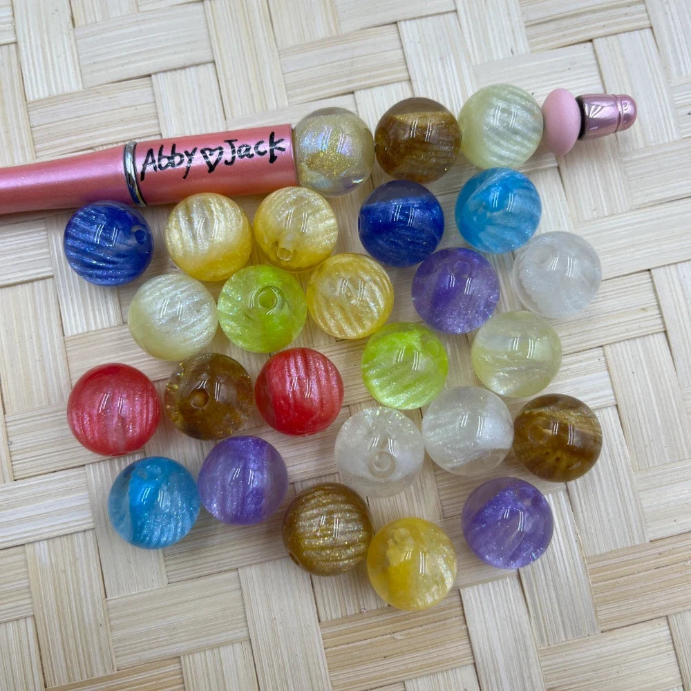 16MM mixed color shimmer galaxy acrylic beads in blue, red, purple, yellow, green, brown, and clear next to a beadable pen on a wicker surface.